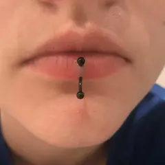Osoba z pionowym kolczykiem w wardze (vertical labret piercing). Czarny, błyszczący kolczyk przechodzi przez środek dolnej wargi.