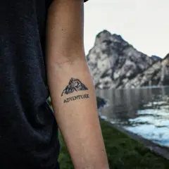 Tatuaż góry z napisem "ADVENTURE" na przedramieniu. W tle jezioro i skaliste szczyty.