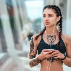 Kobieta z warkoczami i słuchawkami na uszach, trzymająca telefon. Widoczny tatuaż między piersiami przedstawiający kota.
