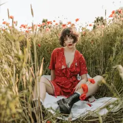 Kobieta w czerwonej sukience siedzi pośród pola maków. Jej tatuaż maki jest subtelnie widoczny.