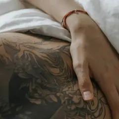 Dłoń na wytatuowanym udzie, jak zdjąć second skina? Delikatne linie tatuażu i biała pościel.