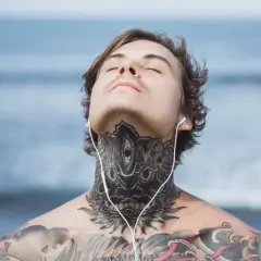 Mężczyzna z tatuażem na obojczyku i szyi, słuchający muzyki na plaży.