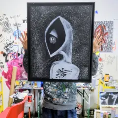 Osoba trzyma obraz z postacią w kapturze, przypominającą styl tatuażu graffiti. W tle ściany pokryte rysunkami i farbami.