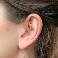 Złoty kolczyk w uchu, ozdobiony cyrkoniami, stanowi subtelny akcent. To modny rook piercing, który dodaje charakteru.