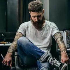 Mężczyzna z brodą i tatuażem męskim na ramionach siedzi w fotelu, trzymając nożyczki.
