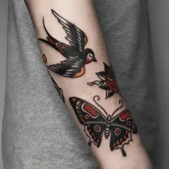 Kolorowy tatuaż tradycyjny na przedramieniu: lecąca jaskółka, motyl i róża.