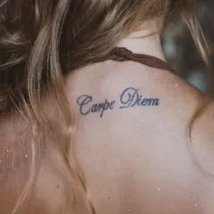 Tatuaże z napisami na karku: "Carpe Diem" na skórze, otoczone jasnymi włosami i drobnym piaskiem.