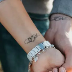 Dwa nadgarstki splecione w uścisku, każdy z subtelnym tatuażem. Na jednym widoczny jest symbol nieskończoności, na drugim ozdobny wzór.