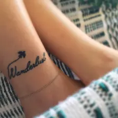 Delikatny tatuaż podróżniczy na nadgarstku: napis "Wanderlust" z samolotem i kotwicą.