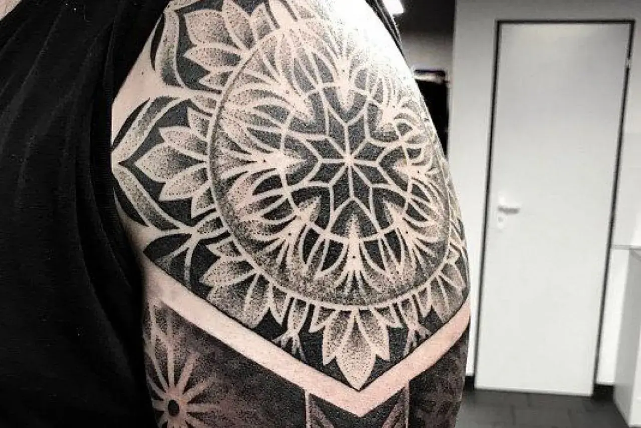 Elegancki tatuaż mandala damski na ramieniu, wykonany techniką dotwork. Wzór przypomina kwiat lub słońce.