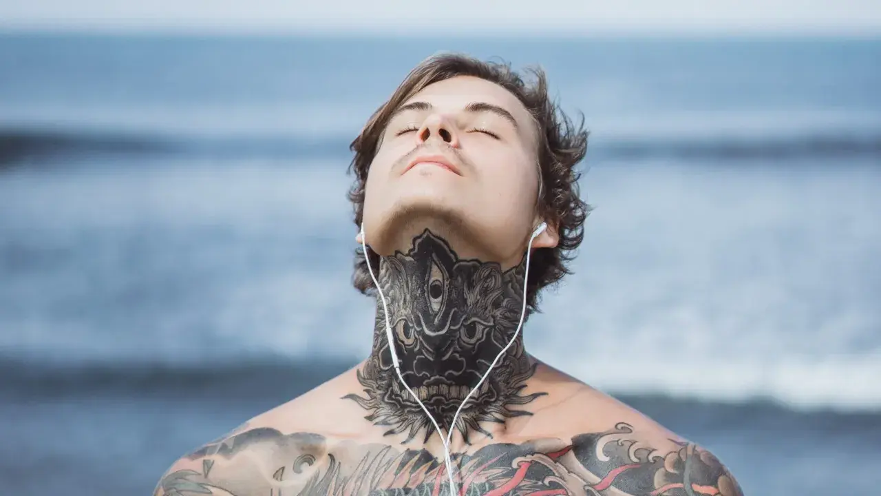 Mężczyzna z tatuażem na obojczyku i szyi, słuchający muzyki na plaży.