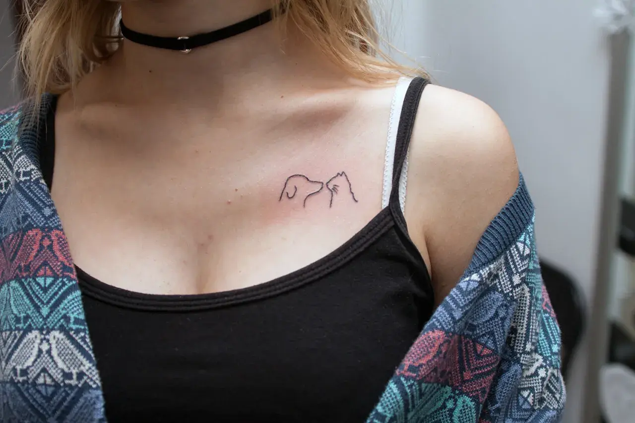Minimalistyczny tatuaż pies i kot na dekolcie, symbol przyjaźni między gatunkami.