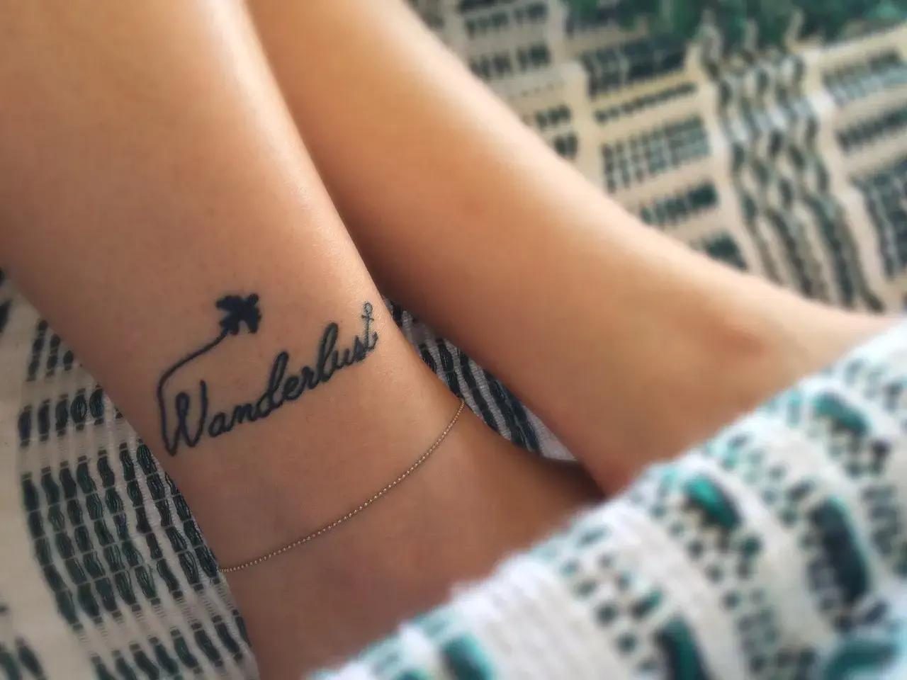 Delikatny tatuaż podróżniczy na nadgarstku: napis "Wanderlust" z samolotem i kotwicą.