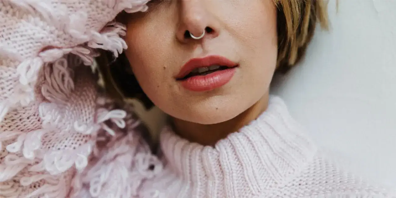 Delikatny różowy sweter otula szyję, a na nosie błyszczy subtelny septum piercing.