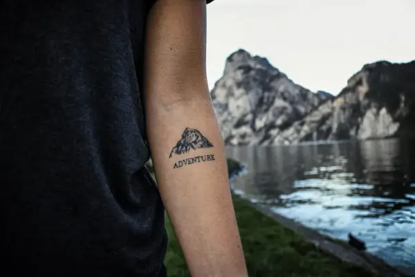 Tatuaż góry z napisem "ADVENTURE" na przedramieniu. W tle jezioro i skaliste szczyty.
